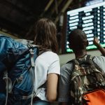 Two backpackers checking the digital board at a train station for travel information.