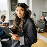 Confident woman with headset and clipboard in modern office environment with colleagues.