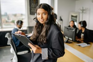 Confident woman with headset and clipboard in modern office environment with colleagues.